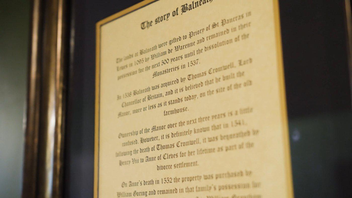 A framed history of Balneath, lettered in black on gold, hanging in the manor